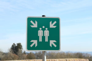 A muster point sign with the classic green background color and four arrows pointing to three figures assembled in the middle.