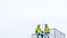 Two workers on an elevated platform, wearing hi-vis safety vests, hard hats, and fall protection harnesses.