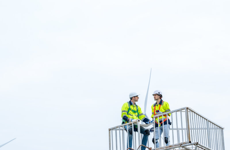 Two workers on an elevated platform, wearing hi-vis safety vests, hard hats, and fall protection harnesses.