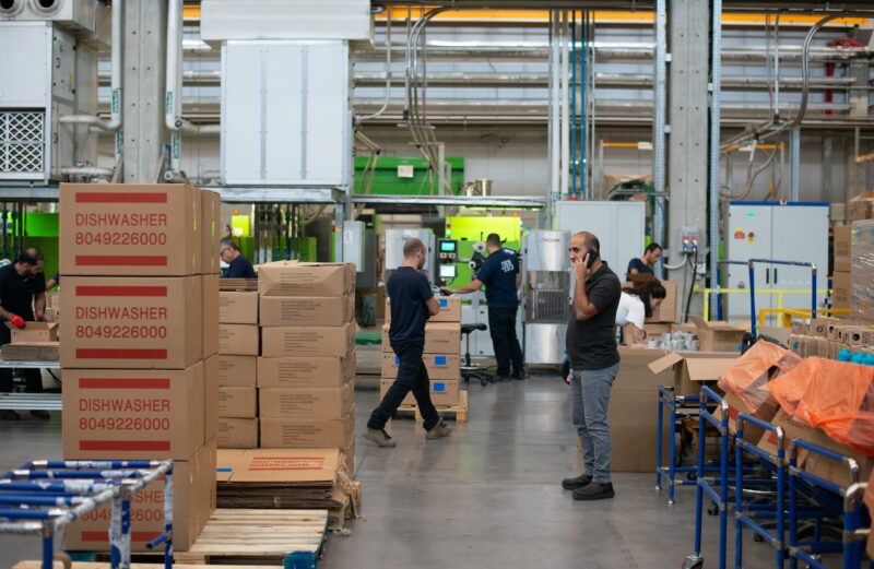 Manager on factory floor, taking a call on a cellphone while employees are working in the background.