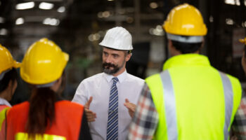 Staff member in hard hat leading a meeting in an industrial setting.