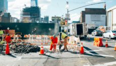 Construction workers doing excavation work on a city street, with a construction site in the background, pedestrians walking by, and cars on the street.