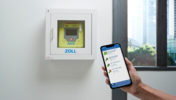 Image depicts a hand holding a smartphone in front of a wall-mounted AED.