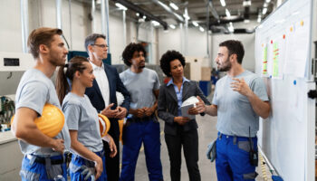 A group of workers gathered for a meeting by a whiteboard in a factory setting.